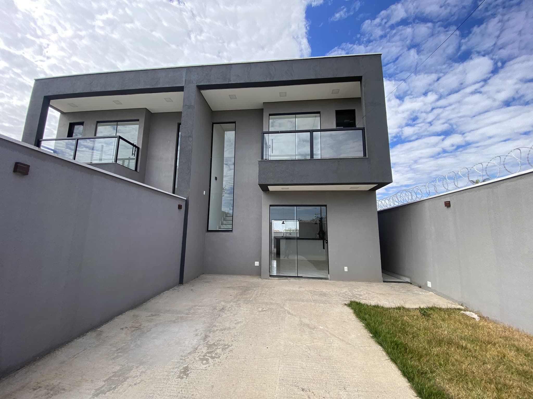 Fachada da residência vista do recuo, com dois pavimentos, varanda com guarda-corpo de vidro e acesso em porcelanato.
